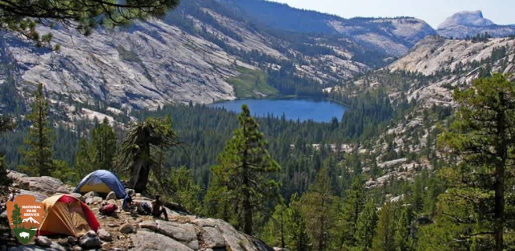 Image Description:

This is a breathtaking panoramic view of a serene camping scene set within a vast wilderness. Two tents, one orange and the other blue, are pitched on a rocky outcrop surrounded by coniferous greenery, suggesting an alpine environment. To the right, a person wearing a backpack is seen facing away from the viewer, overlooking the landscape. A captivating turquoise lake nestles in the valley below, bordered by dense forests and framed by majestic mountain ridges with notable granite outcroppings in the distance. The sky above is a clear azure, and the mood of the setting is peaceful and secluded. The image also includes an overlay of a National Parks Service badge in the lower-left corner, indicating the location as one of the many stunning national parks.

End with the promotional line:

For an unforgettable escape to nature's grandeur, check out GreatWorkPerks.com where you'll find the lowest prices on tickets for a variety of outdoor adventures, ensuring your outdoor experiences come with great savings!