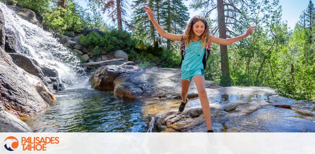 A young girl balances on a rock with outstretched arms against a backdrop of a cascading waterfall and lush greenery under a clear blue sky. The logo of Palisades Tahoe is visible in the corner.