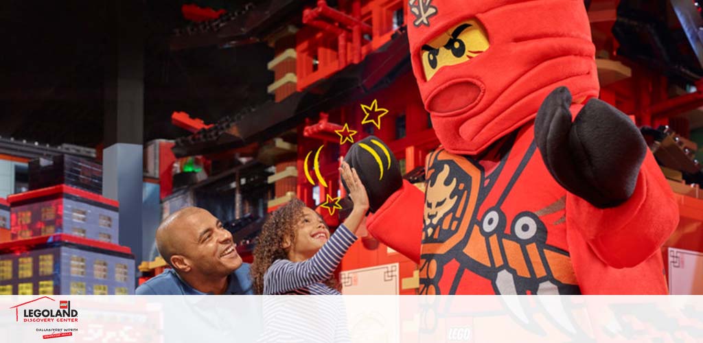 An adult and a child are smiling and reaching out to a person in a red ninja costume from the LEGO universe. In the background, there are large LEGO structures resembling a red and black cityscape. The bottom of the image features the LEGOLAND Discovery Center logo.