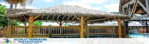 Image depicts Schlitterbahn Waterpark featuring a tropical-themed structure with a thatched roof, surrounded by lush greenery. A clear blue sky with fluffy clouds is visible above, hinting at a sunny day perfect for water activities.