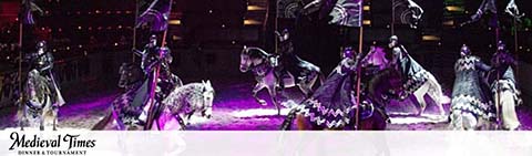 Image shows a Medieval Times dinner and tournament scene. Knights in ornate armor ride on horseback, holding flags, with a dimly lit audience in the background. The arena features theatrical lighting that enhances the dramatic medieval ambiance. The Medieval Times logo is visible at the bottom.