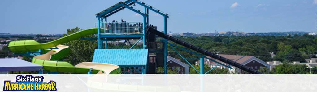 Image of a water slide at Six Flags Hurricane Harbor. The slide features a towering structure with an enclosed green slide spiraling downwards. Below is a lush, tree-filled landscape with a backdrop of a blue sky. In the foreground, a logo is present.