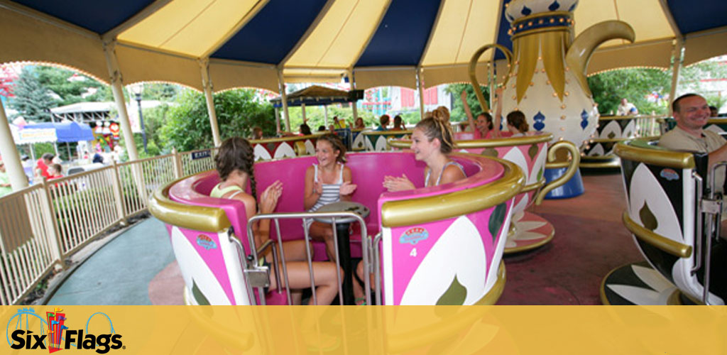 Visitors enjoy a spinning teacup ride at Six Flags. The ride features colorful oversized teacups adorned with pink floral designs, under a yellow canopy. People are seated inside the cups, laughing and chatting as the ride spins. The backdrop includes trees and part of the amusement park. The Six Flags logo is displayed prominently in the corner.