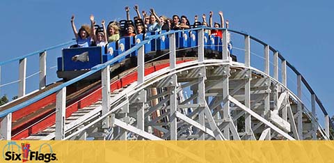 Enjoy exhilarating excitement at Six Flags on a wooden roller coaster. People aboard with arms raised in joy are descending a thrilling drop under a clear blue sky. The Six Flags logo prominently displayed signifies the adventure that awaits.