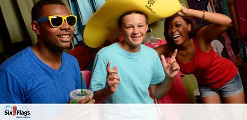 Three friends are enjoying a fun moment together indoors. One wears a giant yellow foam hat and blue shirt, another sports blue sunglasses and holds a beverage, and the third in a red top is posing playfully. A Six Flags logo is visible, suggesting an amusement park setting.