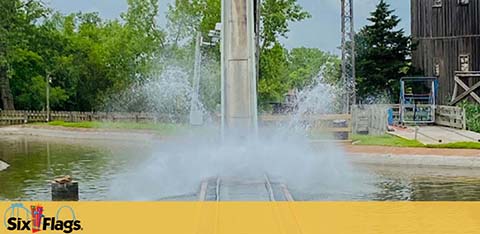 Image shows a splashdown moment at a theme park ride, where a water ride car creates a large spray as it hits the water. In the background, a wooden structure suggests the ride's theme. The Six Flags logo is visible, indicating the park's brand.