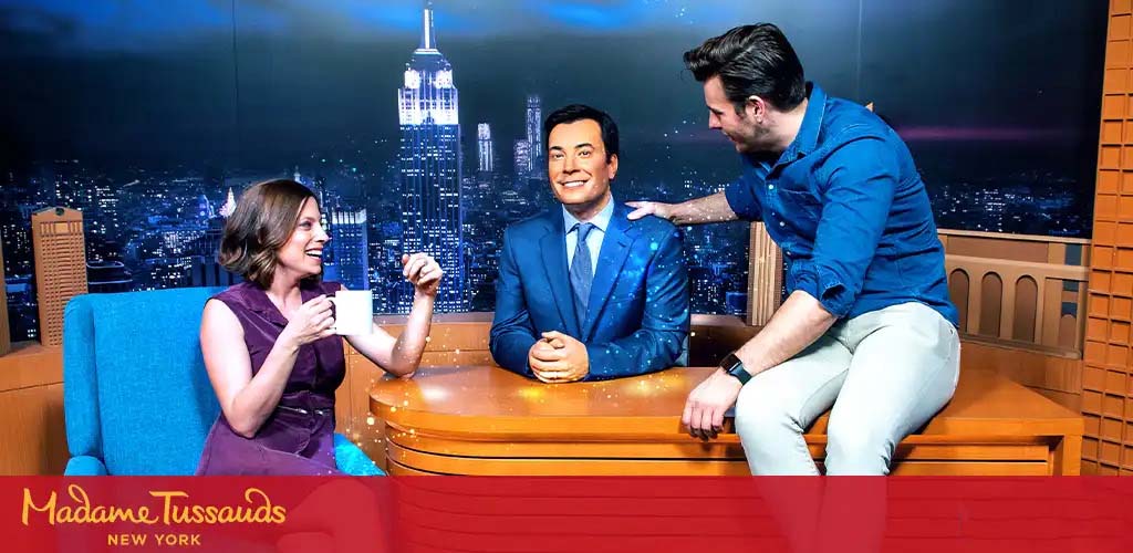 Image shows a person seated on a talk show set at Madame Tussauds New York, engaging with a wax figure resembling a television host. They are both behind a desk with the nighttime NYC skyline backdrop. The visitor is gesturing towards the wax figure with a friendly demeanor.