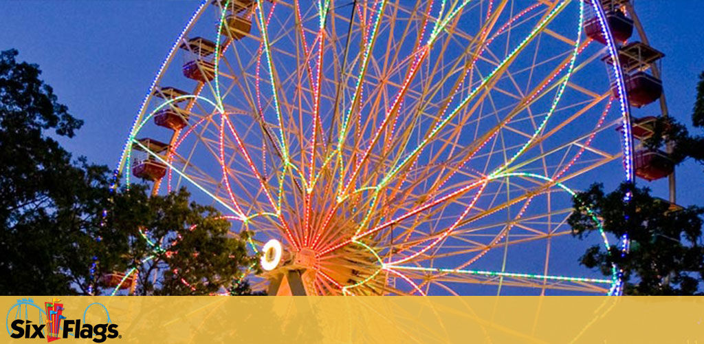 Image shows a brightly lit Ferris wheel at dusk with colorful, neon lights along its structure against a dimming sky. Trees are partially visible in the background. The Six Flags logo is present in the upper portion of the image, suggesting the location is a Six Flags amusement park.