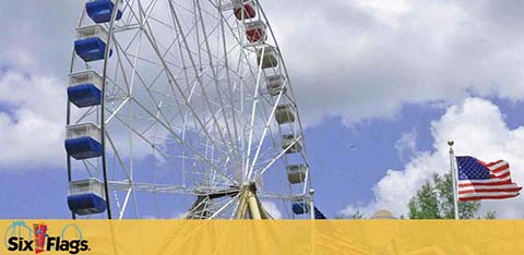 Image shows a large Ferris wheel with blue and white capsules against a partly cloudy sky. In the background, an American flag flutters in the breeze. In the foreground, a logo for Six Flags is displayed above a yellow banner.