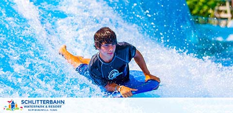 A person is bodyboarding on a simulated wave at Schlitterbahn Waterpark and Resort. They are focusing intently on navigating the rushing water. Bright sunshine enhances the vibrant blue of the wave. The logo of Schlitterbahn is visible in the corner of the image.