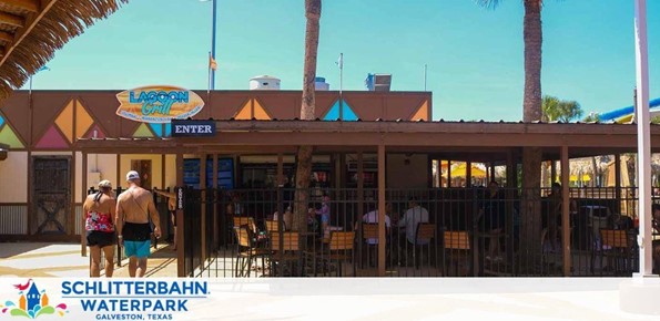 Image shows the entrance to Lagoon Grill at Schlitterbahn Waterpark in Galveston, Texas, under a clear blue sky. People are seen walking towards the brown building with vibrant signage, surrounded by palm trees suggesting a warm, tropical setting.