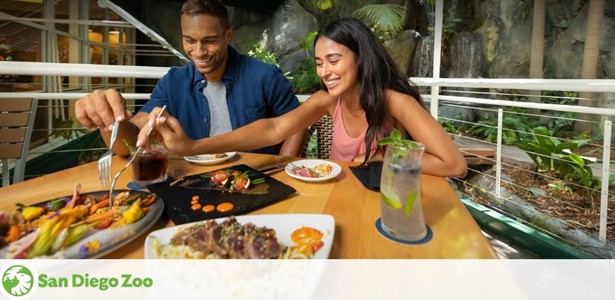 Image shows a joyful couple dining at the San Diego Zoo. They're casually dressed, sharing a meal with a variety of colorful dishes on the table, surrounded by lush greenery indicative of the zoo's tropical atmosphere. The San Diego Zoo logo is visible in the corner.