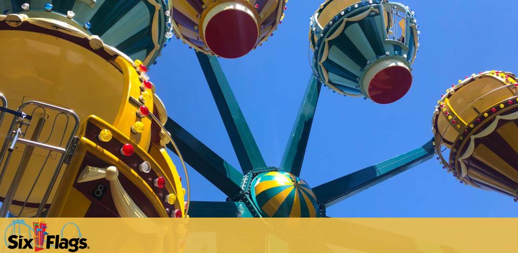 A vibrant amusement park ride with yellow, green, and blue colors against a clear blue sky. Below the logo for Six Flags, colorful swinging gondolas are captured from a ground perspective, looking up as they dangle and await riders.