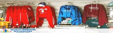 Image displaying a lineup of colorful branded merchandise on a shelf at Schlitterbahn Waterpark. From left to right, there are multiple red hoodies with white text, a red hoodie with a lifeguard design, blue tie-dye hoodies, and green and maroon hoodies with white text. The backdrop features various waterpark gear.