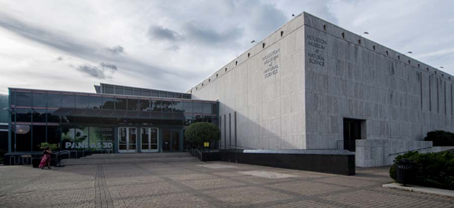 Image of a modern building with a prominent sign indicating it is a 3D film theater. The theater entrance features glass doors flanked by posters and a couple is sitting nearby. The adjacent building is clad in textured concrete with the museum's title etched on its surface. Clouds suggest a partly cloudy sky above.