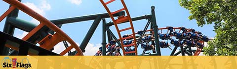 Image shows a roller coaster with orange tracks and blue cars, filled with riders in mid-descent. Lush green trees in the background, under a clear blue sky. The Six Flags logo is visible in the bottom left corner, signaling the amusement park brand.