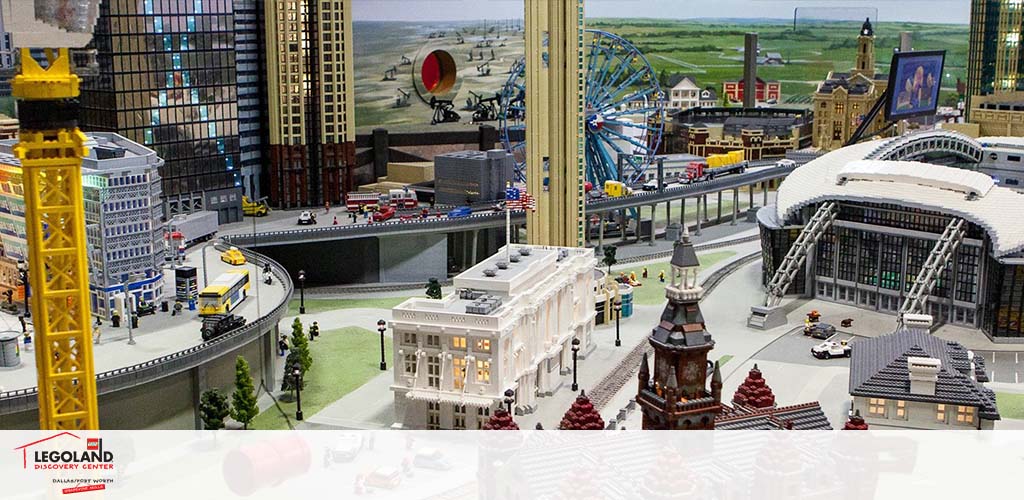 Image features a Legoland Discovery Center display. Visible is a detailed miniature Lego city with buildings, a Ferris wheel, roads with vehicles, and a train. Structures resemble a bustling urban center with a stadium-like venue to the right. The Legoland logo is in the bottom left corner.