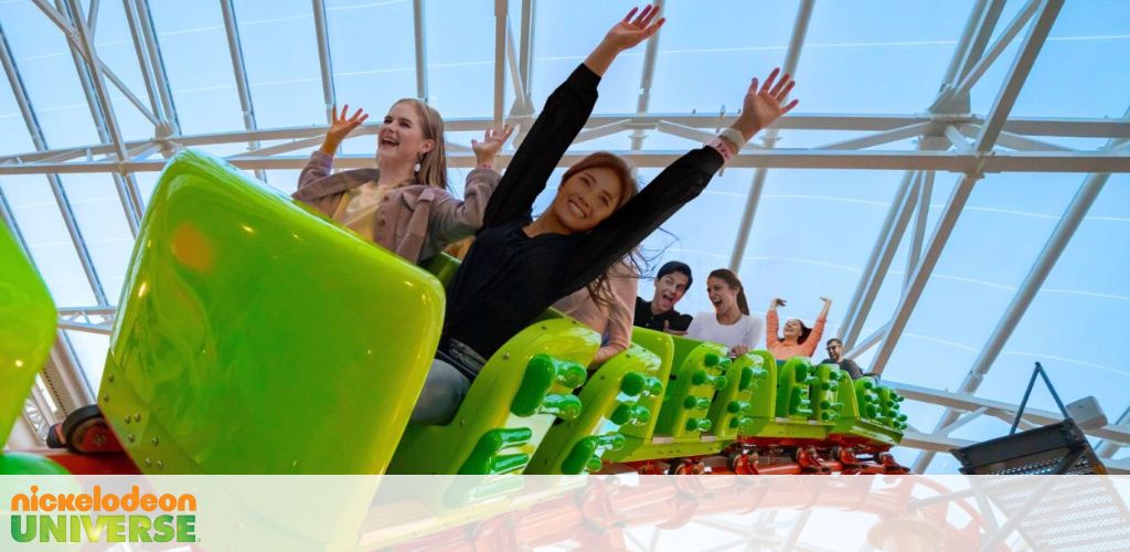 Image shows people with raised hands enjoying a roller coaster ride at Nickelodeon Universe. The excitement is evident in their expressions. The coaster car is vibrant green under a large glass structure, suggesting an indoor amusement park setting.