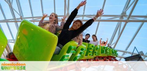 Image shows people aboard a bright green roller coaster under a translucent ceiling, arms raised in excitement. In the foreground, a person is smiling towards the camera. The backdrop features the text 'Nickelodeon Universe'. Emotion is evident by the joyous expressions.