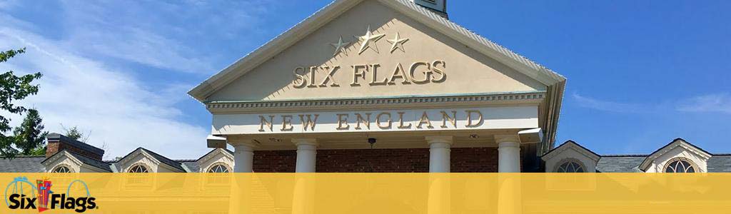 An image displaying the entrance facade of Six Flags New England. The photo shows the name and logo prominently featured on a classical-style building with a gabled roof and ornamental details under a clear blue sky.