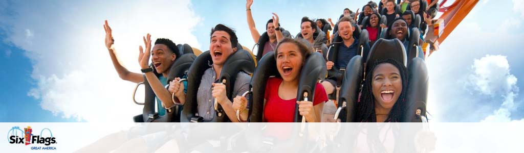 Excited visitors enjoy a thrilling roller coaster ride against a clear blue sky at Six Flags Great America. They are seated in a row, hands raised in joy, with expressions of exhilaration and delight. The Six Flags logo is visible at the bottom left.