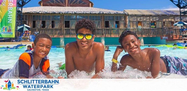 Three joyful children are leaning on the edge of a pool with waves, at Schlitterbahn Waterpark in Galveston, Texas. The sunny sky reflects on water's surface, and a wooden building with flags is in the background. They're wearing swimwear and one has sunglasses.