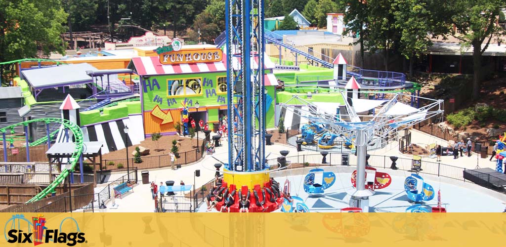 Image shows an amusement park with colorful rides and attractions. In the foreground, there's a blue spinning ride with red seats elevated above a platform. The background features a roller coaster with green tracks, and a 'Funhouse' with vividly colored walls and playful designs. The Six Flags logo is visible, indicating the park's brand. The sky is clear and the setting appears lively and entertaining.