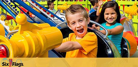 Image shows two joyful children riding a colorful amusement park ride. A boy in the foreground is in an orange shirt, smiling widely with excitement. A girl behind him, wearing a purple shirt, is also enjoying the ride. Both seem to be having a fun experience. The Six Flags logo is visible in the lower part of the image, indicating the location.