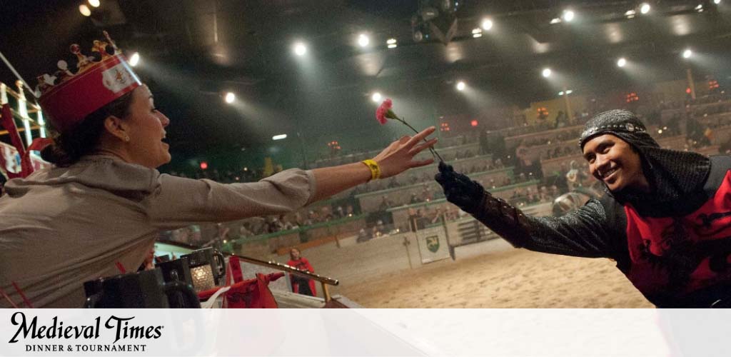 A performer in medieval attire hands a flower to a smiling audience member wearing a crown at a Medieval Times dinner and tournament event. The arena in the background is dimly lit with spectators watching the interaction.