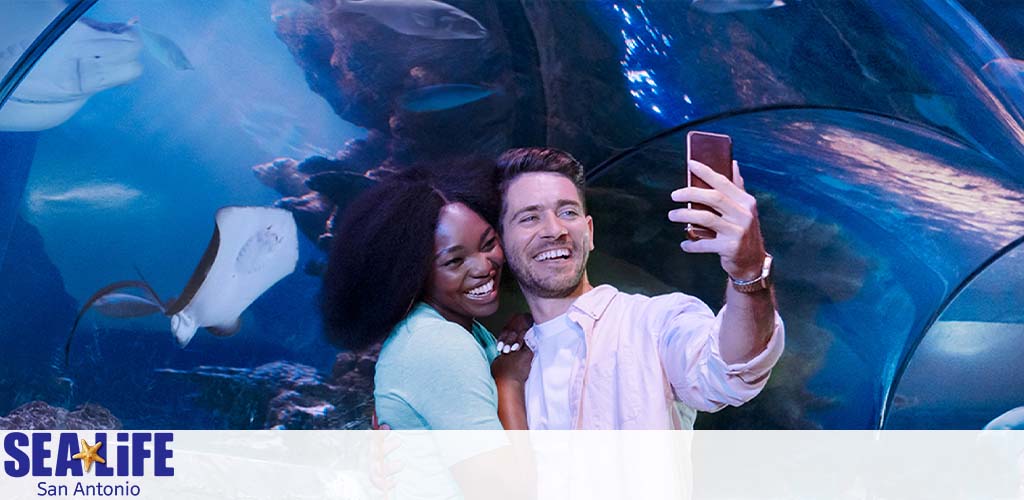 A joyful couple takes a selfie at SEA LIFE San Antonio. They're smiling, surrounded by an underwater scene with rays and fish in a large blue-lit aquarium. The SEA LIFE logo is visible at the bottom left.