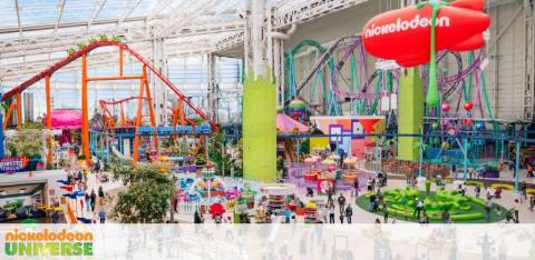 Inside Nickelodeon Universe, an indoor theme park, showcasing colorful roller coasters, attractions, and seating areas. The space is covered by a translucent ceiling allowing natural light in, with vibrant green pillars and a large Nickelodeon balloon upfront.