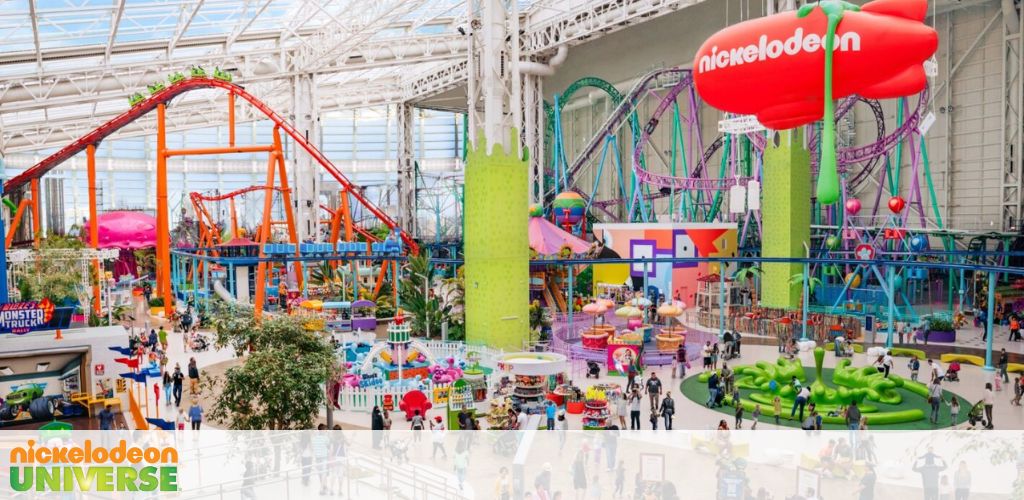 Image of Nickelodeon Universe, an indoor amusement park. Brightly colored roller coasters twist above a lively crowd. Attractions and themed areas with various activities offer entertainment, highlighted by the signature green slime design. Large windows illuminate the space with natural light.