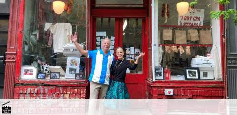 Image of a man and woman standing with raised hands in a friendly wave outside a vibrant red storefront. The window displays several framed pictures and signs, suggesting a gallery or shop with a creative flair. The building features traditional detailing and lanterns that add to its welcoming charm.