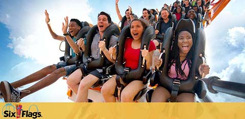 Image features a diverse group of excited riders on a roller coaster. They are mid-ride with expressions of thrill and joy under a blue sky. The Six Flags logo is visible, indicating the theme park.