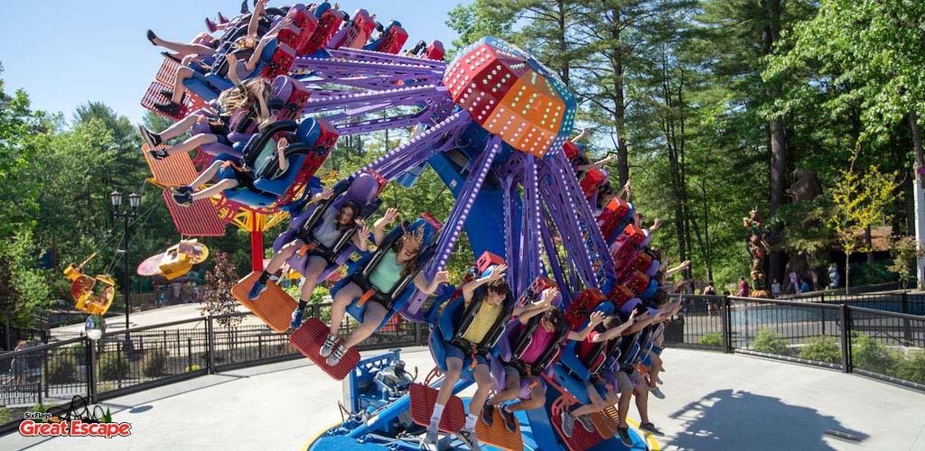 Visitors enjoy a thrilling ride at an amusement park on a sunny day. The ride features multiple arms with attached seats, spinning and flipping people through the air. Trees surround the area, adding a natural backdrop to the scene. Riders appear ecstatic amid the excitement.