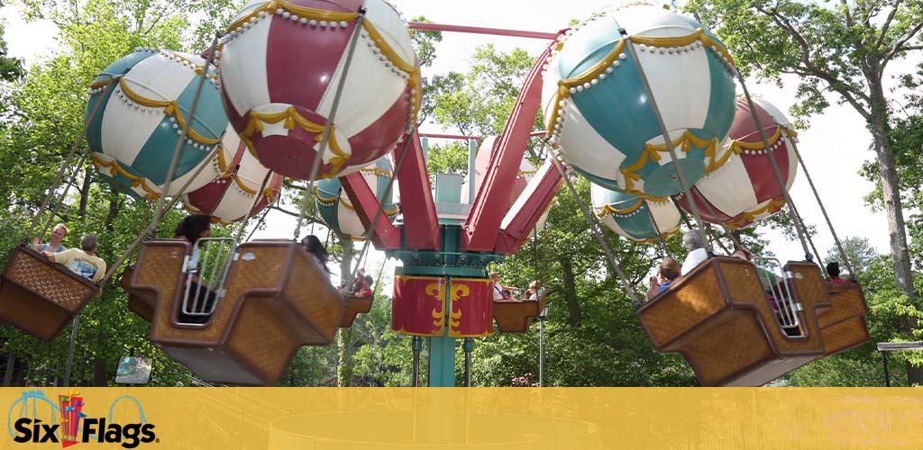 Image of a balloon-themed ride at Six Flags amusement park. Visitors are seated in gondolas designed to resemble hot air balloons with colorful patterns, suspended from a central structure, against a backdrop of green trees. The Six Flags logo is visible on the image.