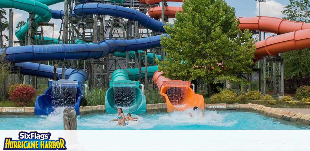 Image of a water park at Six Flags Hurricane Harbor showing a colorful array of intertwined water slides in blue, teal, and orange, ending in splash pools. People are seen enjoying the slide, emerging into the water with the backdrop of green trees under a partly cloudy sky.