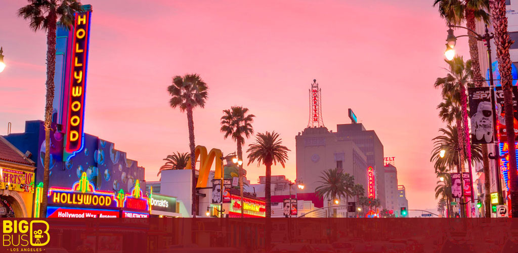 This image captures the vibrant scene of Hollywood Boulevard at dusk. The sky is awash with the warm hues of pink and orange, providing a striking backdrop to the scene. Prominent in the foreground is the brightly lit neon sign of the Hollywood Wax Museum, glowing with electric blues, yellows, and reds. Flanking the museum are palm trees and various other colorful neon signs, including one for the fast-food chain McDonald's. In the middle distance, additional illuminated signage and billboards can be seen, with a notable vertical sign that reads 'Hollywood,' adding to the iconic streetscape. The legendary El Capitan Theatre appears as well, marked by a neon tower with its name at the top. Sprinkled throughout the image, street lamps cast a soft glow that reflects on the buildings below. The famous setting is bustling with culture and the electric energy characteristic of Los Angeles. 

At FunEx.com, we provide unbeatable discounts and the lowest prices on tickets to the most sought-after destinations, including the star-studded streets of Hollywood.
