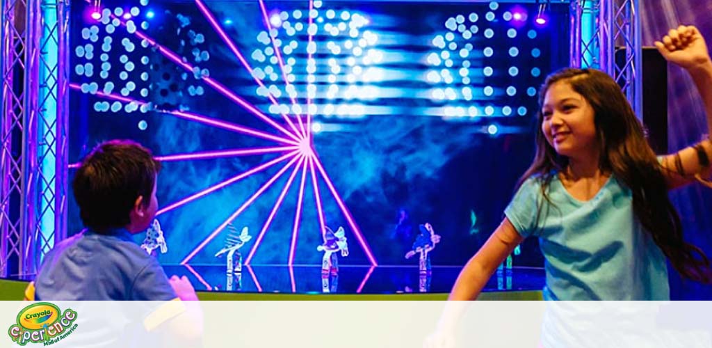 A boy and a girl are enjoying a lively show with a neon-lit stage featuring animated figures. The girl, on the right, is smiling and dancing with her arm raised, while the boy, on the left, watches the performance intently. Bright pink beams of light and blue stage lights add to the festive atmosphere.