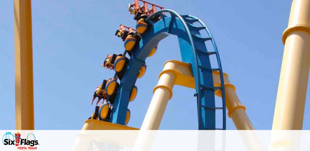 Image of a blue roller coaster with riders upside down at the crest of a loop. Yellow beams support the structure against a clear sky. The Six Flags Fiesta Texas logo is visible, indicating the location of the amusement ride.