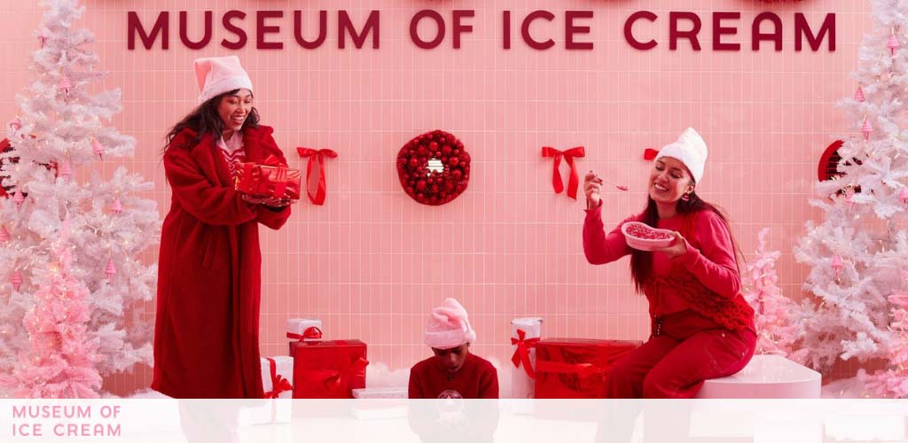 Image shows a holiday-themed display at the Museum of Ice Cream with two people in red winter clothing against a backdrop of pink walls and white Christmas trees. One person is holding a gift and the other is enjoying a bowl of ice cream.