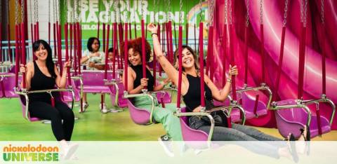 Three people are seated on a swing ride at Nickelodeon Universe, smiling with their thumbs up. Bright pink and green decor enhances the playful atmosphere with the slogan 'Use Your Imagination' in the background.