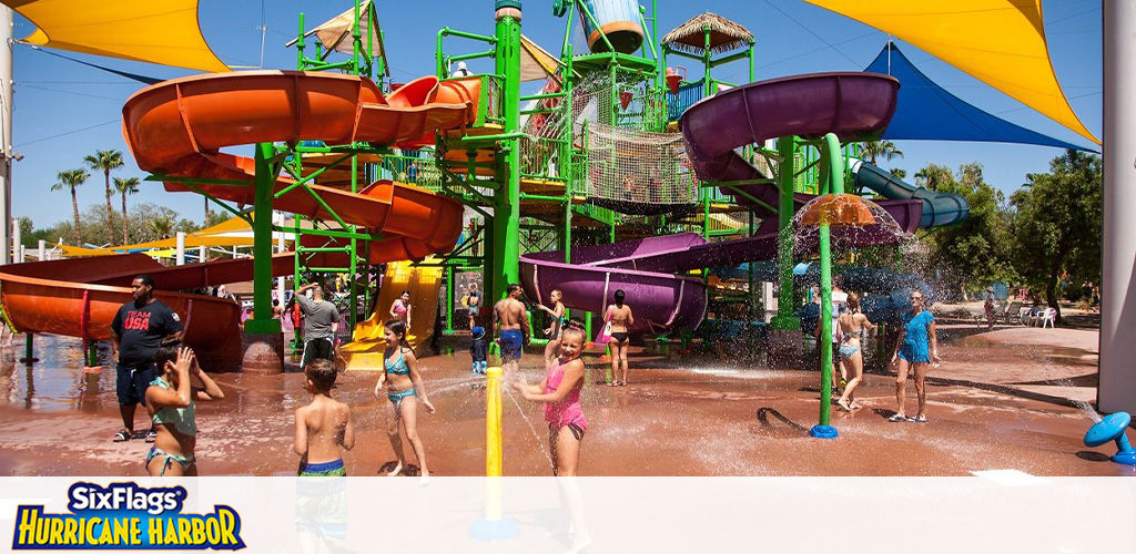 Image shows a colorful water park scene at Six Flags Hurricane Harbor. People of various ages are enjoying sunny weather, splashing in shallow water, and standing near a giant water play structure with multiple twisting slides in orange and purple, shaded by a large yellow and blue canvas above.