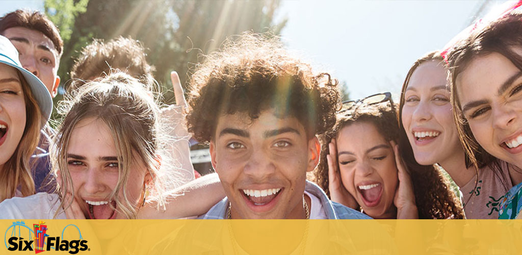 Image shows a group of cheerful young people smiling and posing closely together outdoors. Sunlight filters through, highlighting the Six Flags logo at the top, implying a fun day at the amusement park. Bright expressions and sunny weather contribute to a joyful atmosphere.