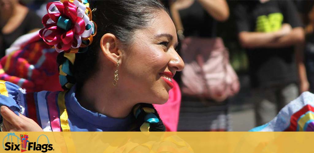 Image shows a woman smiling in profile, wearing a traditional colorful dress with a vibrant, multicolored hair accessory. A blurred crowd is in the background. The Six Flags logo is in the top left corner. Warm tones and joyful atmosphere are depicted.