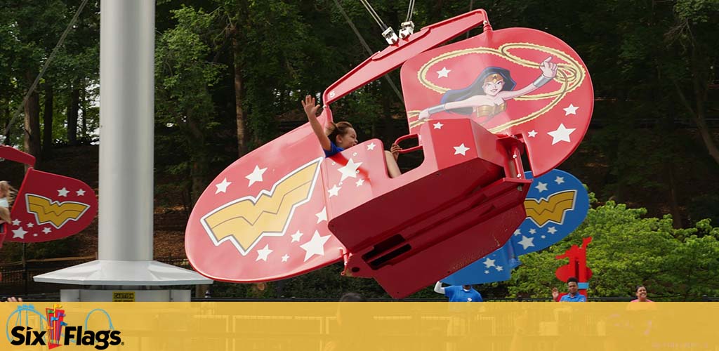 Photo of an amusement park ride at Six Flags with Wonder Woman branding. Riders sit in red, open-air pods adorned with the superhero's iconic symbols, swinging around a central pole. The background has lush green trees and the sky is overcast. A child raises their arm, mimicking flight. The Six Flags logo is visible.