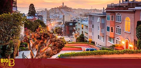 This image features the iconic view of a zigzagging street taken during a beautiful sunset with its warm glow bathing the scene. To the left, a series of trimmed trees and a red-brick pathway lead the eye down the windy road. Elegant residential buildings flank one side of the street, showcasing a variety of architectural styles and warm-colored facades. The road itself is adorned with red pavement and a series of cars lining the curbs. In the background, the rolling cityscape extends into the distance, culminating in a hill with structures populating its side, under a soft blue sky with hints of pink and orange near the horizon.

At FunEx.com, we take pride in offering our customers exclusive discounts and savings so you can enjoy picturesque destinations like this without pushing your budget uphill. Browse through our selection for the lowest prices on tickets to your next adventure!