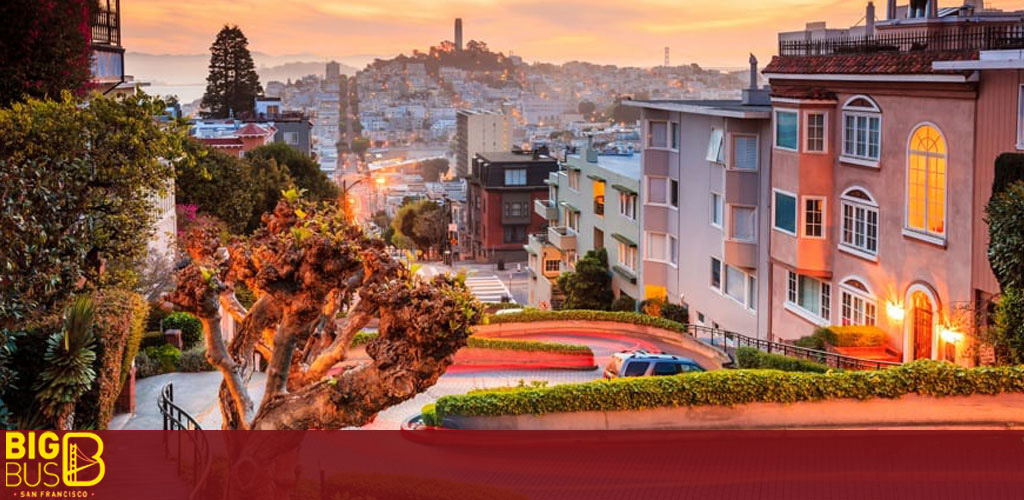 This image presents a scenic view of San Francisco's iconic Lombard Street, known for its steep, one-block section with eight hairpin turns. The photo captures the street at either sunrise or sunset, evidenced by the warm golden hues illuminating the frame. To the left, a series of green foliage and a textured tree add to the charm of the winding red-bricked road, which is lined with headlights and taillights indicating the presence of vehicles. The surrounding architecture displays a variety of building styles with the nearest structure painted in cream and boasting tall, arched windows that emit a soft glow. The cityscape stretches into the background, showcasing a dense aggregation of buildings leading up to a distant hill, possibly Telegraph Hill, crowned by the slender silhouette of Coit Tower. The sky is clear, transitioning from a sunlit amber near the horizon to a deeper blue above, suggesting the calm of early morning or the tranquility of early evening. At FunEx.com, our commitment to providing you with an easy and enjoyable booking experience is matched by our determination to secure the lowest prices, ensuring that your trip to San Francisco comes with both unforgettable memories and unbeatable savings on tickets.