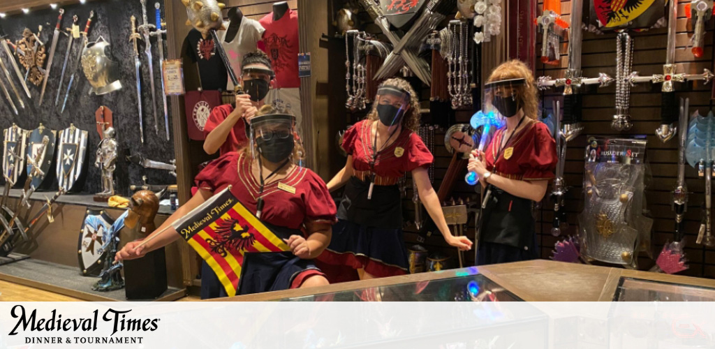 This image features a vibrant and animated scene inside a Medieval Times gift shop where four employees, all wearing face masks, are engaging with potential customers. They are dressed in red and yellow uniforms, giving off a medieval-themed aesthetic. The staff is positioned behind a counter adorned with various novelty items that reflect a chivalric era. The shelves behind them are neatly lined with a collection of medieval memorabilia such as swords, shields, helmets, and other themed merchandise. The atmosphere emits a sense of excitement and historical adventure.

This shop seems to be a place where visitors can immerse themselves in the medieval experience and bring a piece of the past home with them. Remember, at FunEx.com we pride ourselves on offering the lowest prices and greatest savings on tickets to a wide array of attractions and events, ensuring your medieval adventure won't take a joust at your wallet!