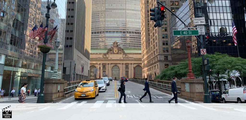 Street view in a city with pedestrians crossing the road at an intersection marked E 40 St. Taxis and cars are present. Tall buildings line the street, and an ornate building with a large clock resides in the background. American flags hang from a lamppost and the building ahead.